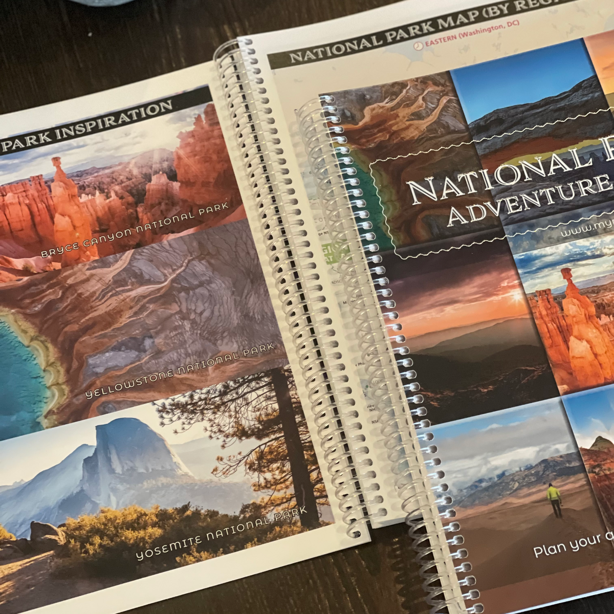 A spiral-bound planner or notebook featuring national park map and inspiration pages, photographed open on a dark wooden tabletop. The cover reads 'National Park Adventure' and shows scenic collage imagery including red rock canyons, mountains, and landscapes.