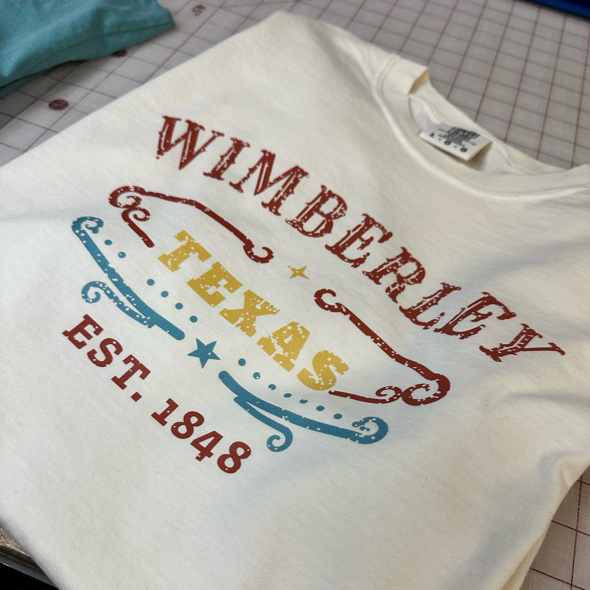 Ivory Comfort Colors tee with Wimberley Texas Est. 1848 design, folded on a cutting mat