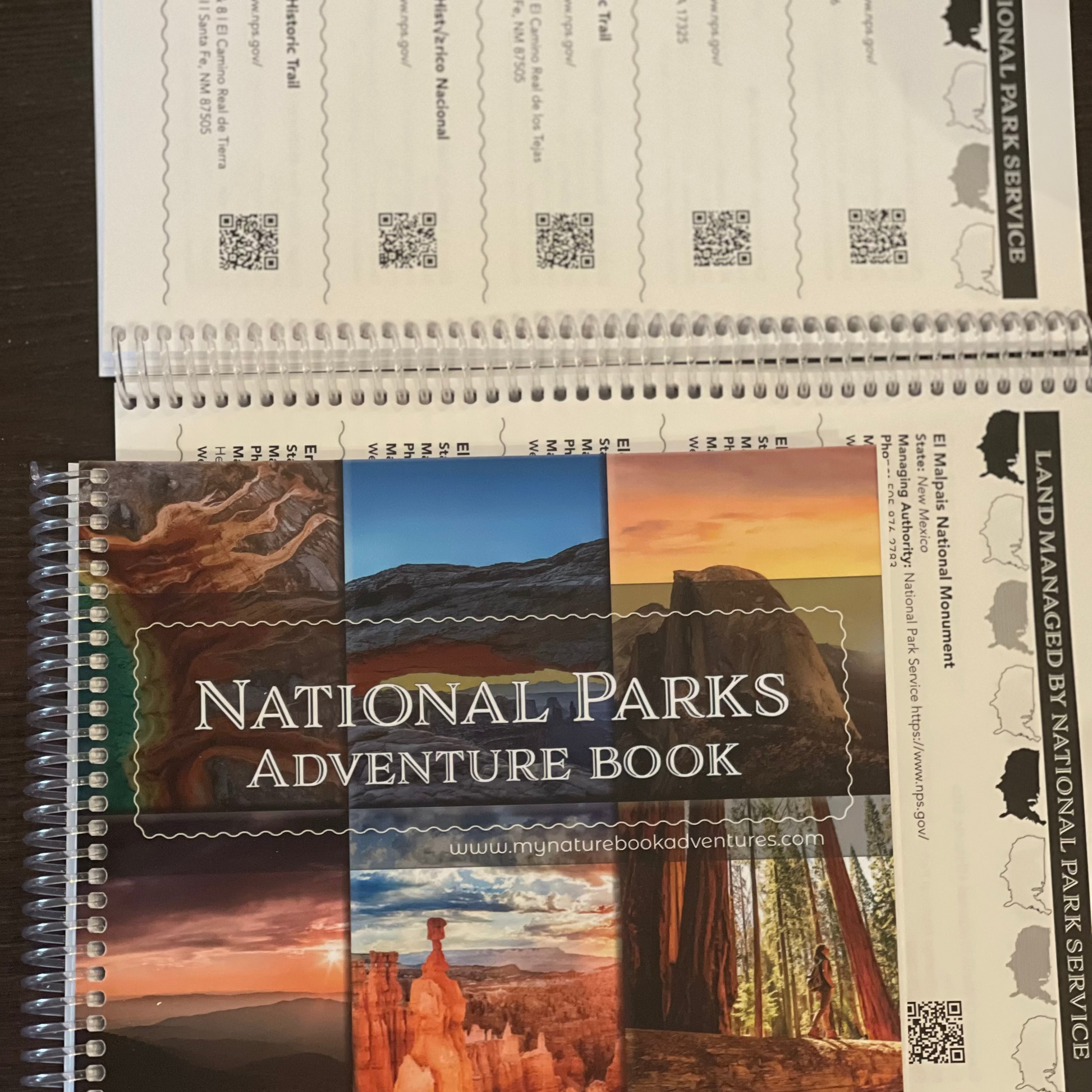A spiral-bound National Parks Adventure Book with a collage cover showing various national park landscapes, placed on top of printed National Park Service forms on a dark wooden surface.