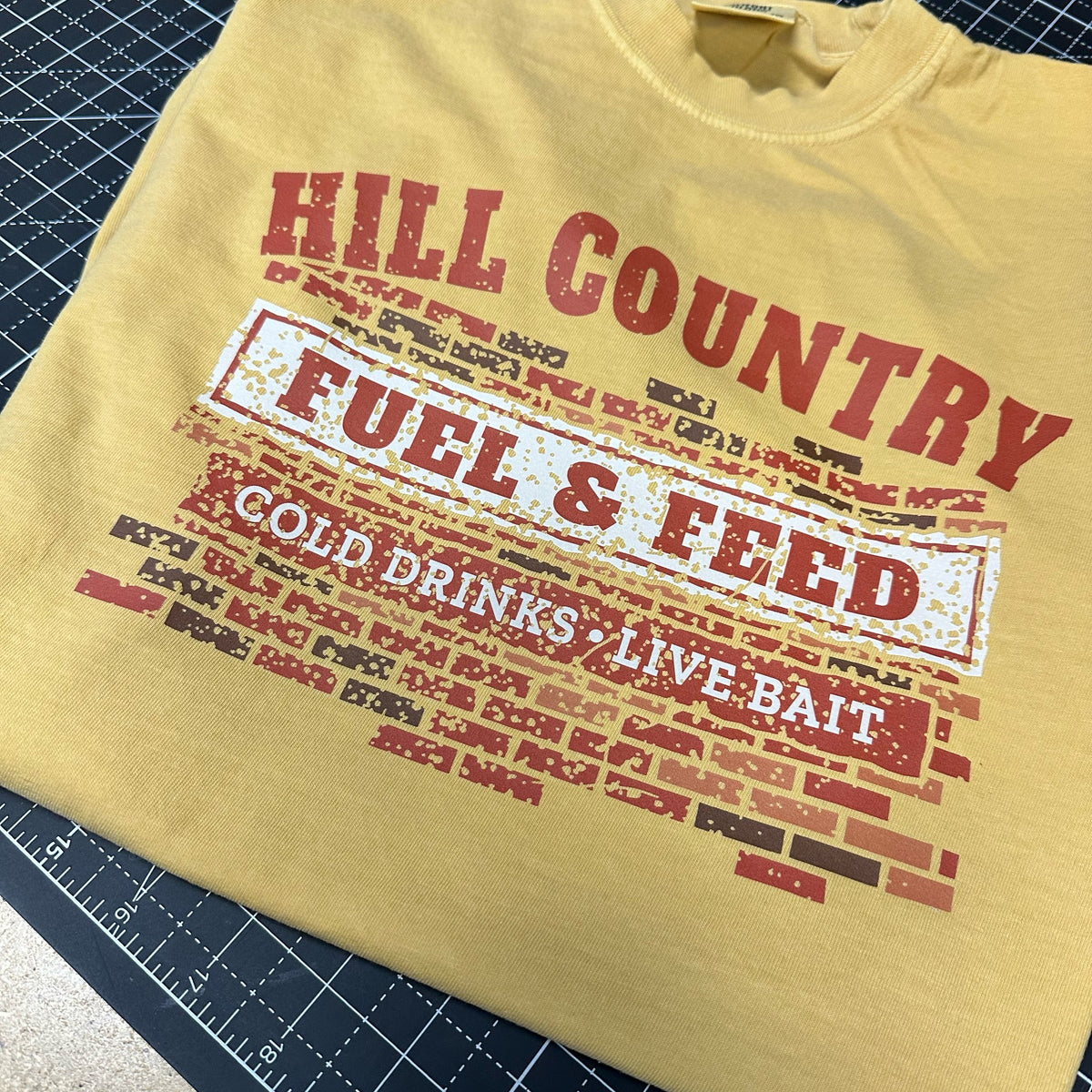Mustard Comfort Colors tee with Hill Country Fuel & Feed, Cold Drinks, Live Bait design, folded on a cutting mat