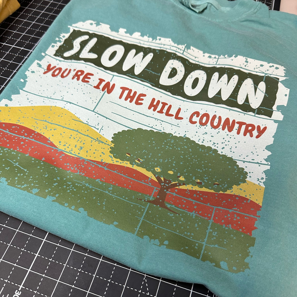 Seafoam Comfort Colors tee with Slow Down, You're in the Hill Country design, folded on a cutting mat